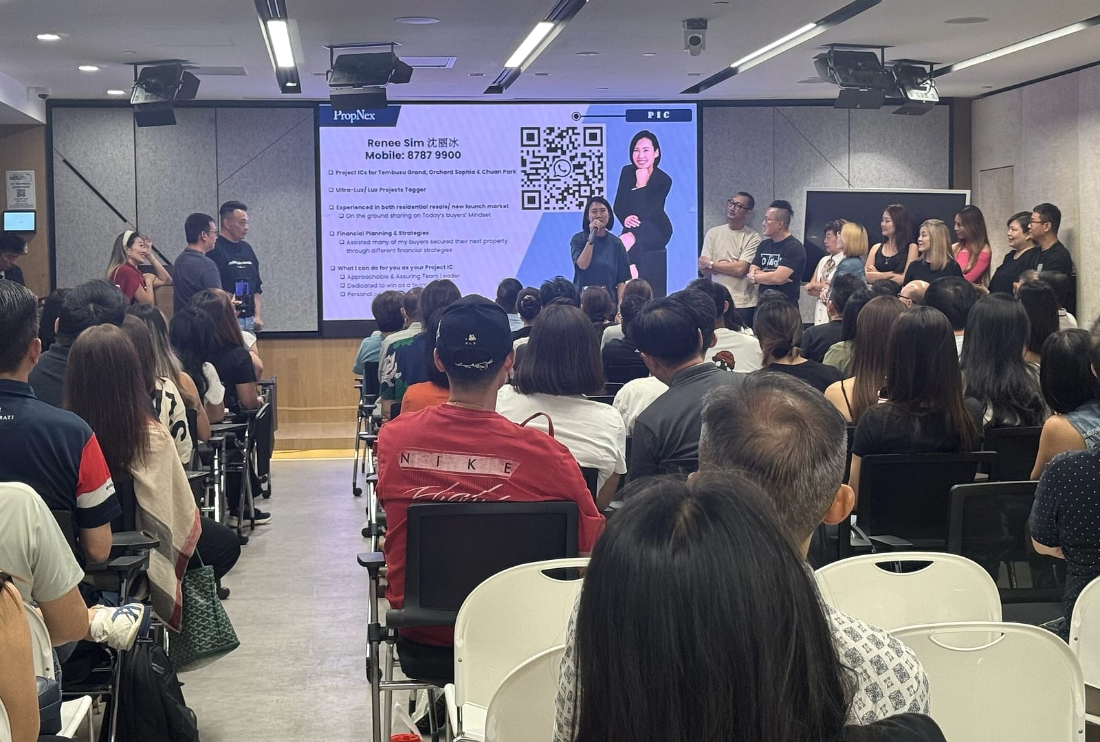 Renee Sim speaking into a microphone at the front of a full PropNex training room, in front of a projected slide listing her project IC roles, with around eighty seated agents facing her.