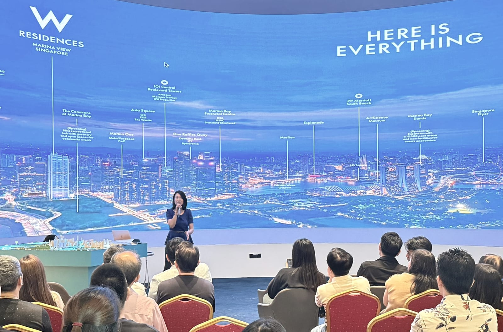 Renee Sim speaking with a microphone in front of a wall-sized Marina Bay skyline render at the W Residences Marina View gallery, addressing a seated audience.